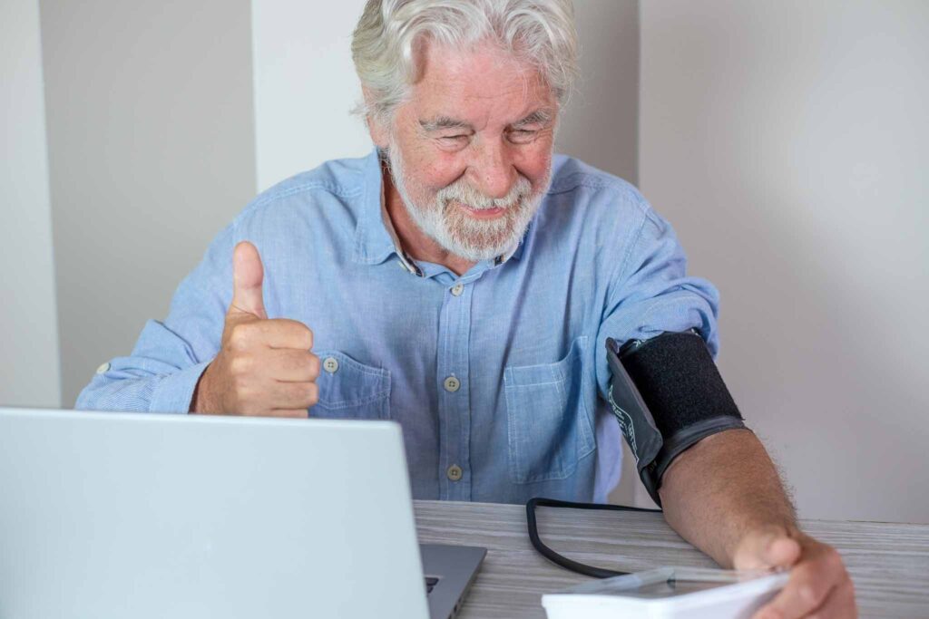 Video call. Bearded senior man suffering from hypertension sitting at home talking remotely with doctor on computer webcam while live checking blood pressure and reporting results to doctor